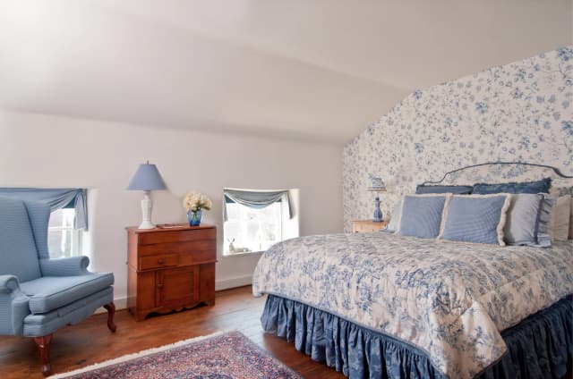 A bedroom with floral blue and white wallpaper, matching bedding, and cushions. Wooden drawer and blue armchair add warmth. Bright natural light.
