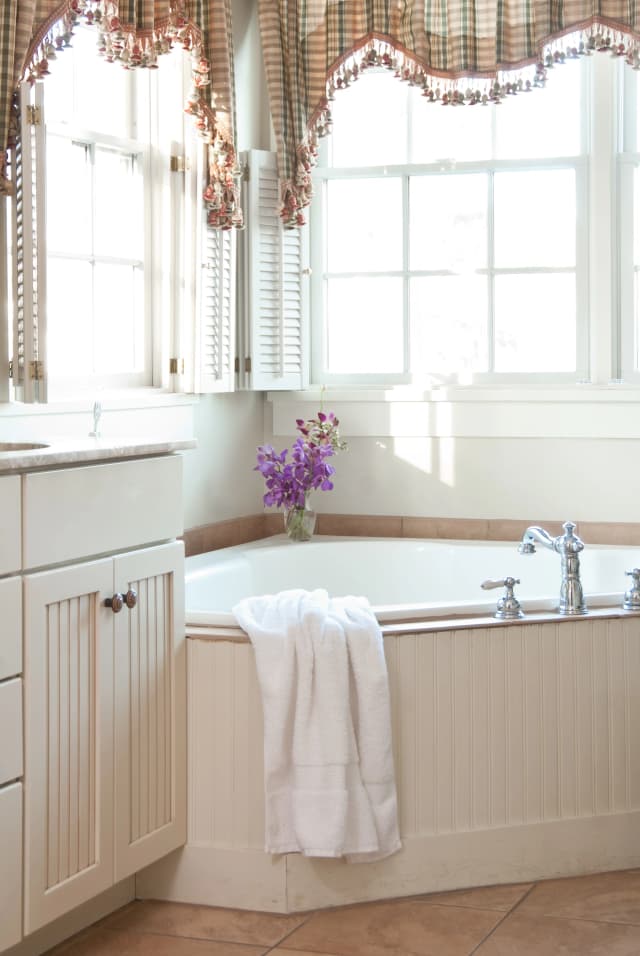 A bright elegant bathroom corner with a white bathtub, a folded white towel draped over the edge, a vase of purple flowers, and large windows with plaid curtains.
