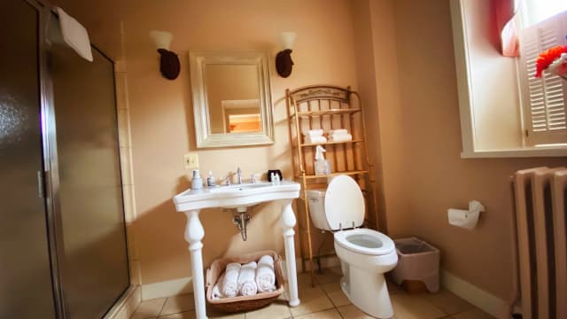 Bathroom with a white sink, open toilet, wooden towel rack, basket of towels, shower stall, window with shutters, and beige walls.