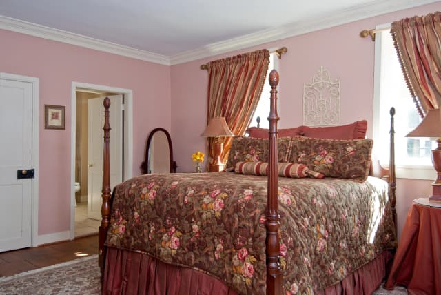 Betty Suite bedroom view from right side: Elegant bedroom with a four-poster bed draped in floral bedding. Pink walls and striped curtains add warmth. A standing mirror and table lamps complete the cozy, classic look.