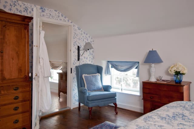Margo Suite, a vintage bedroom with floral wallpaper. Features a blue armchair, wooden furniture, blue lamp, and a small window with a blue curtain. Calming ambiance.