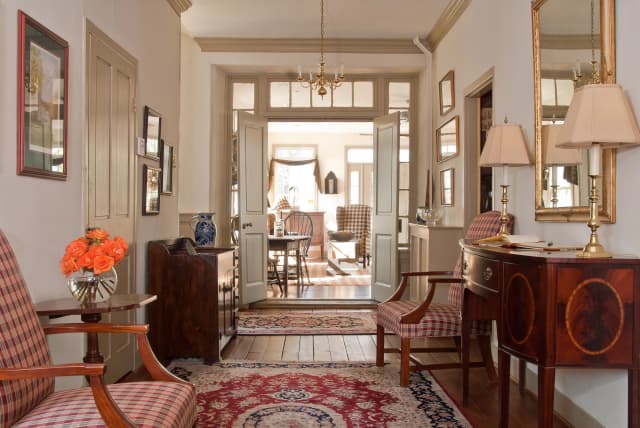 Cozy hallway with vintage decor, featuring plaid chairs, a red-patterned rug, and warm lighting. Open doors reveal a sunlit dining area.