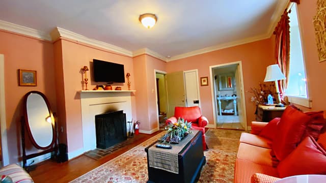 Cozy living room with peach walls, a lit ceiling light, fireplace, and wall-mounted TV. Red sofa and armchair, floral rug, and decorative plants. Warm ambiance.