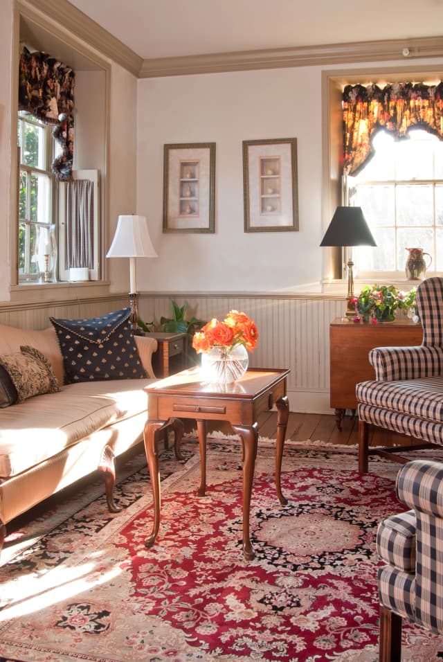 A cozy living room with sunlight streaming through windows. Features include a floral rug, striped armchair, beige sofa, and a wooden table with a vase of orange flowers. Neutral walls and framed art add warmth.