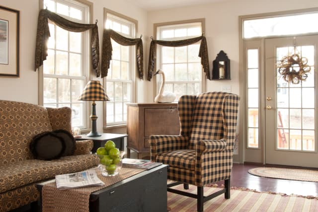 Cozy living room with checkered armchair and patterned sofa. Large windows with elegant drapes brighten the space. A table holds apples and newspapers.