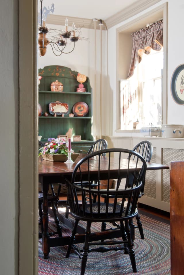 Cozy dining room with a wooden table, four black chairs, and a centerpiece of flowers. A green hutch with decor stands beside a sunlit window.