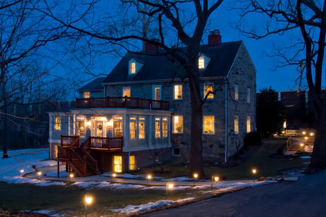 Large stone house at dusk, warmly lit from within. Snow patches and glowing path lights create a cozy, inviting ambiance. Bare winter trees surround.