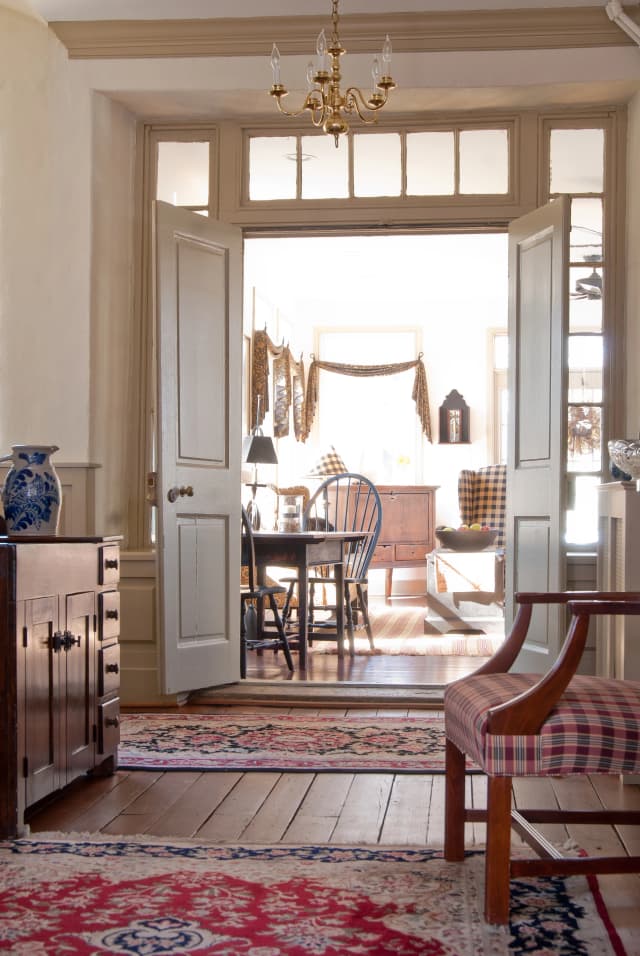Cozy vintage interior features wooden floors, patterned rugs, a blue vase, and checkered chairs. Sunlit dining area with a chandelier and open doors.